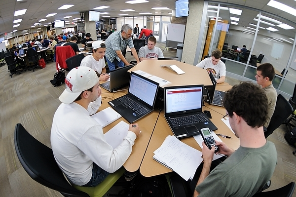 A teacher answers student questions at a hexagonal table with students working together on problem solving using computers in one of the CEETE Program Active Learning Classrooms.